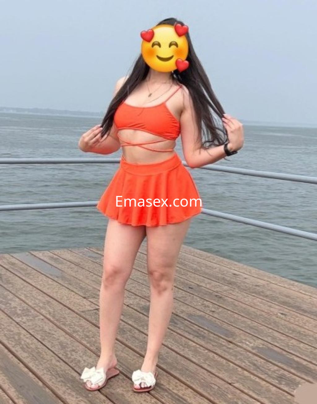Escort en Valladolid, con cabello largo y suelto, viste un conjunto de bikini naranja y una falda corta, posando junto al agua.
