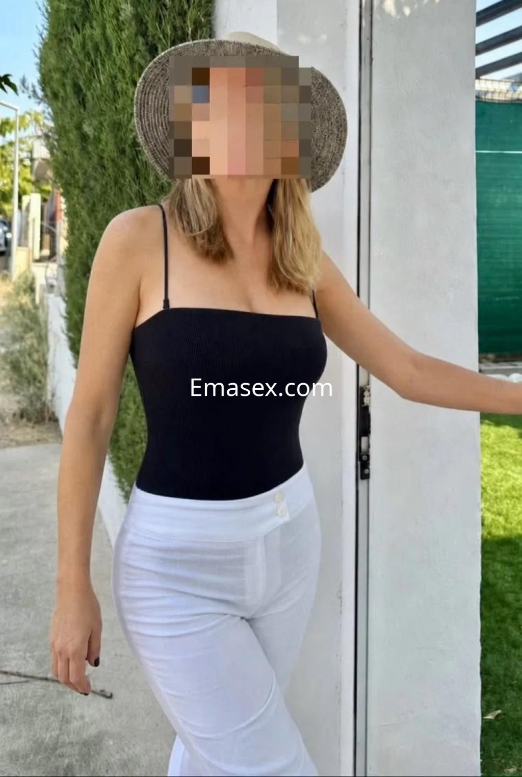 Puta en Zaragoza luciendo un top negro sin mangas y pantalones blancos, de pie junto a una pared blanca.