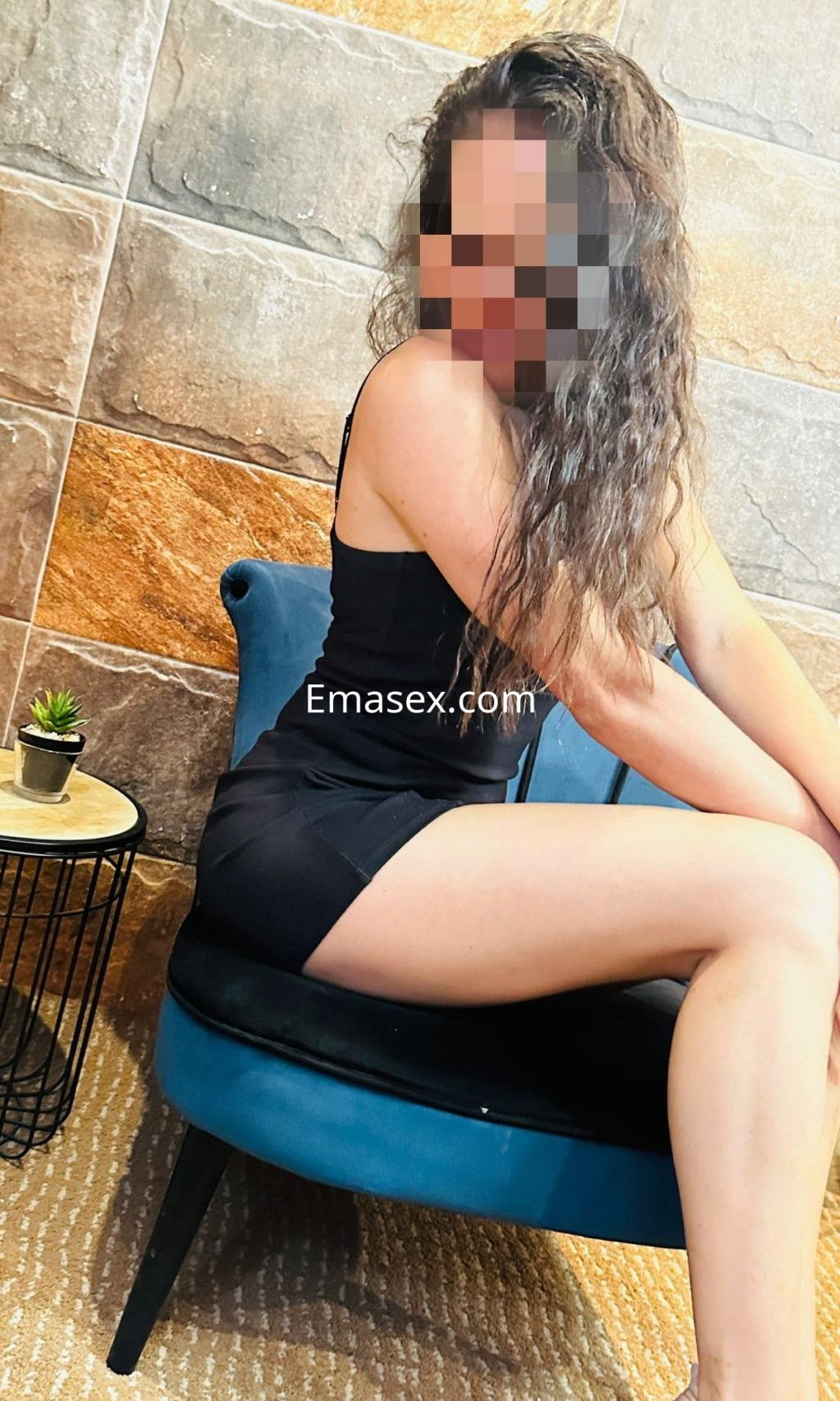 Escort sentada en un sillón azul en Zaragoza, luciendo un vestido negro ajustado y con cabello rizado.