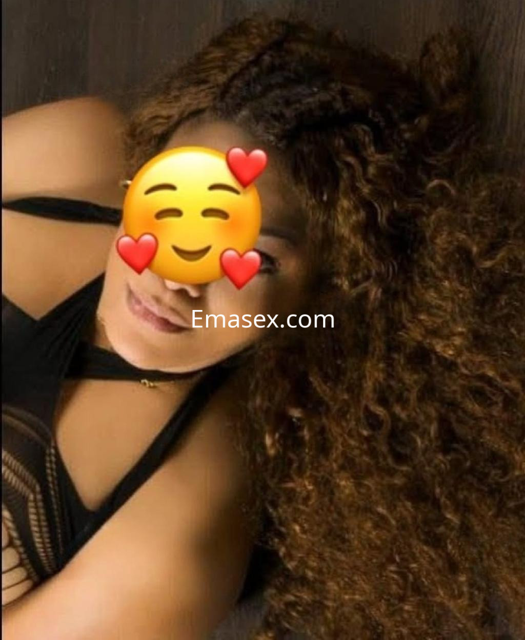 La puta descansa sobre un fondo oscuro, luciendo un atuendo negro con detalles que resaltan su figura y cabello rizado.