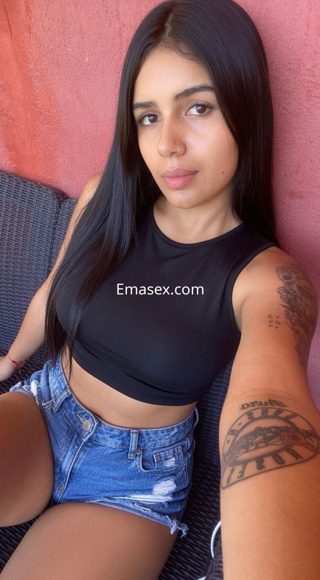 Escort en Sevilla con cabello largo y lacio, viste una camiseta negra y shorts de mezclilla, posando sonriente.