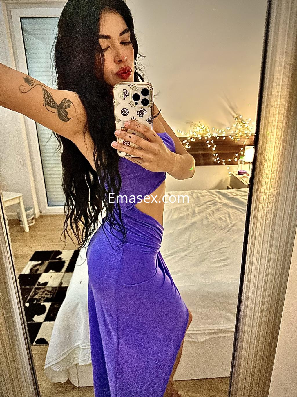 Puta de pie frente a un espejo, con vestido largo morado que resalta su figura y cabello largo y ondulado.