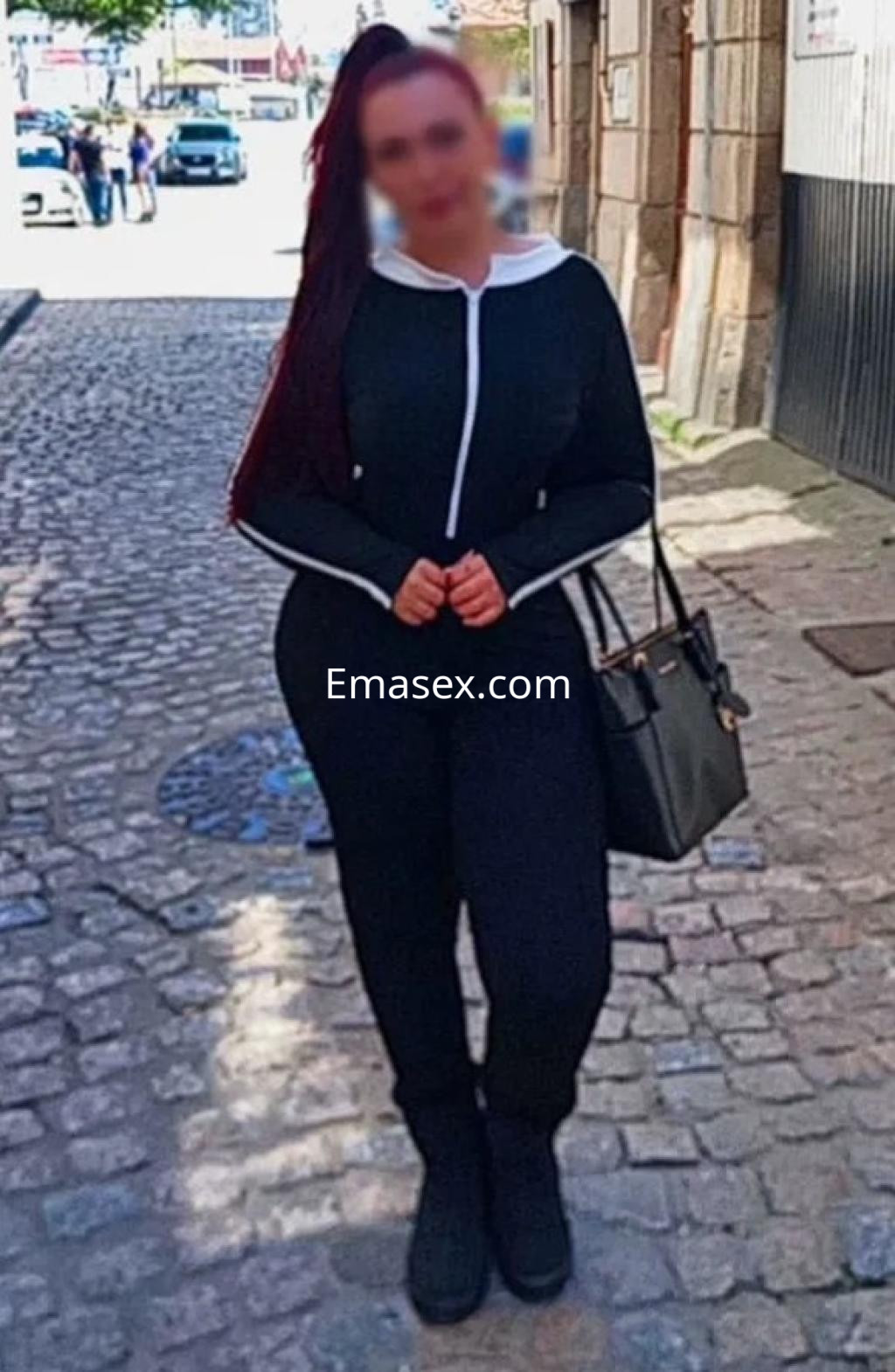 Puta con cabello largo y rojo, vestida con un conjunto negro ajustado y bolso, posando en una calle de Vigo.