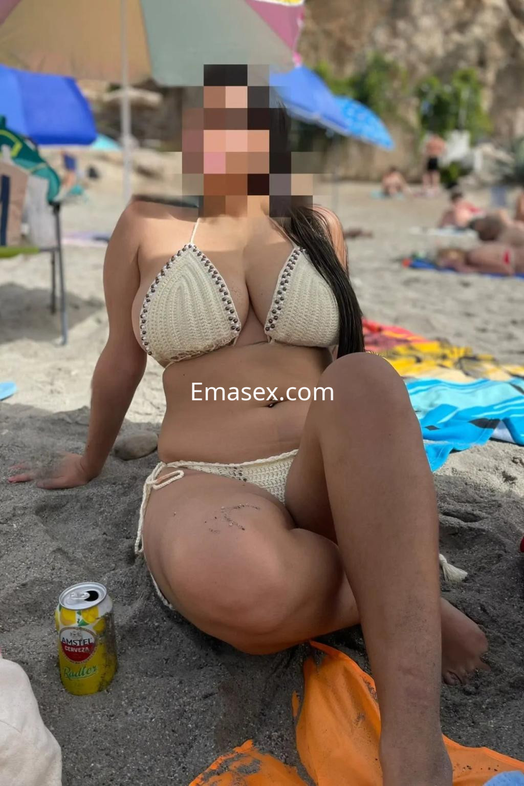 Escort en Córdoba posando en la playa con un bikini de crochet, sentada sobre la arena con una bebida a su lado.
