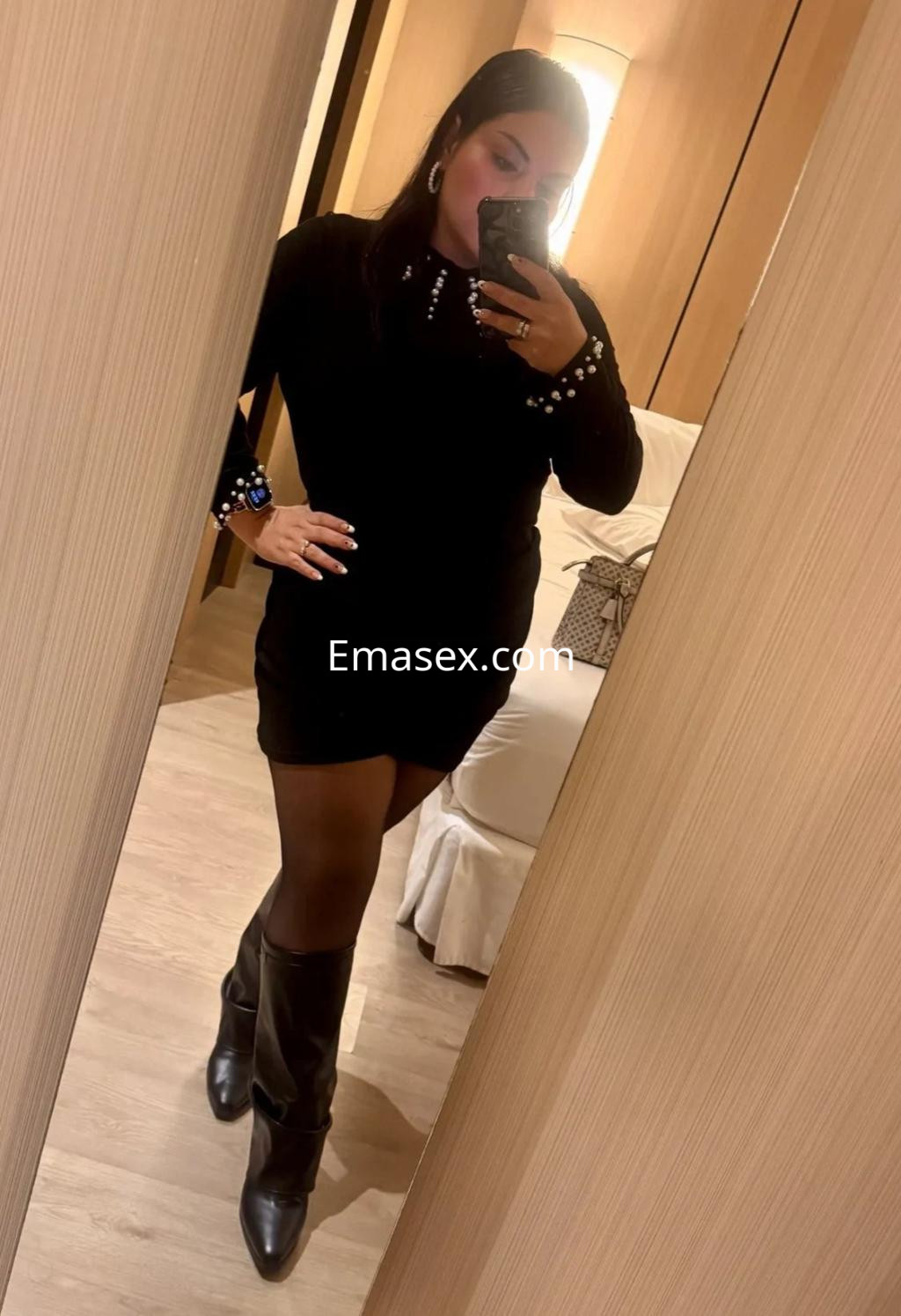 La puta viste un vestido negro ajustado con detalles brillantes y botas altas, posando frente al espejo en Córdoba.