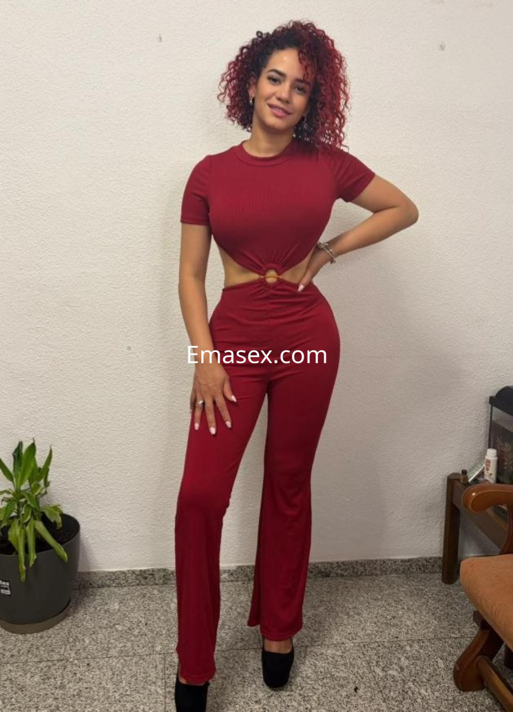 Escort en Córdoba, luciendo un conjunto rojo con cut-out y tacones, posando con una sonrisa y cabello rizado.
