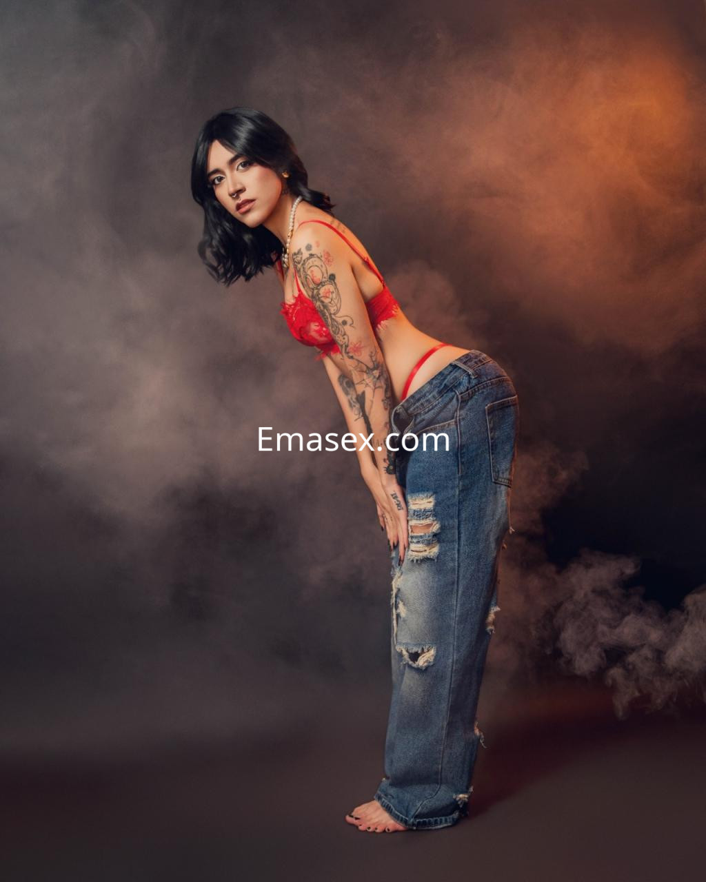 Puta con cabello oscuro y ondulado, viste un sujetador rojo y jeans desgastados, posando en un fondo con humo.