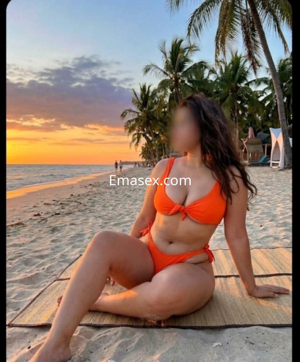 Escort en Ourense, sentada en la playa al atardecer, vistiendo un bikini naranja y rodeada de palmeras.