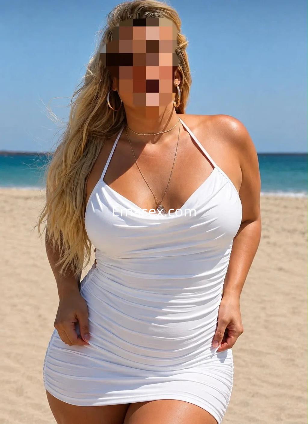 Escort en Oviedo con vestido blanco ajustado, cabello largo y ondulado, posando en la playa junto al mar.