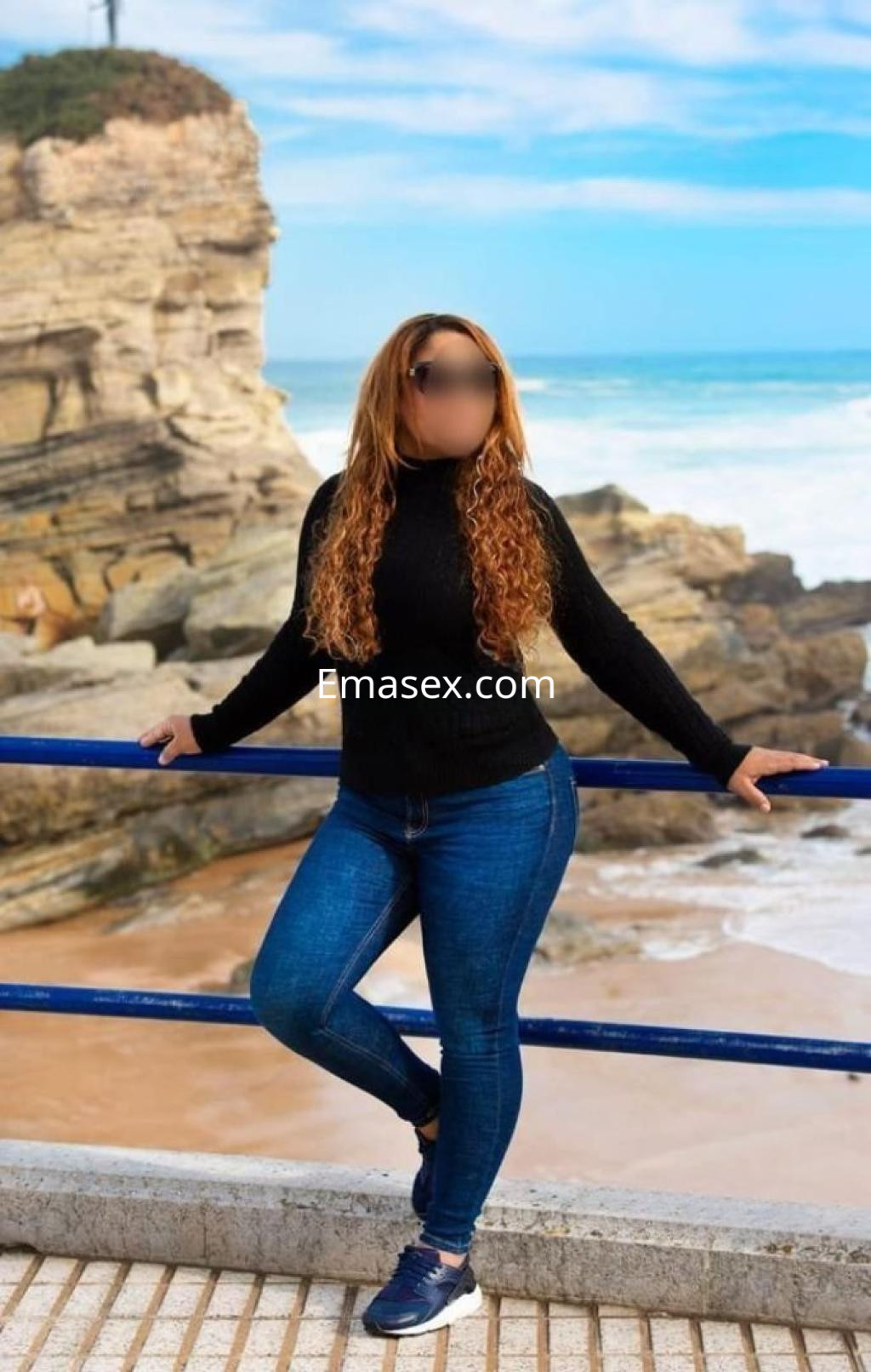 Escort posando en un entorno costero de Zaragoza, con cabello rizado y atuendo casual de suéter negro y jeans.