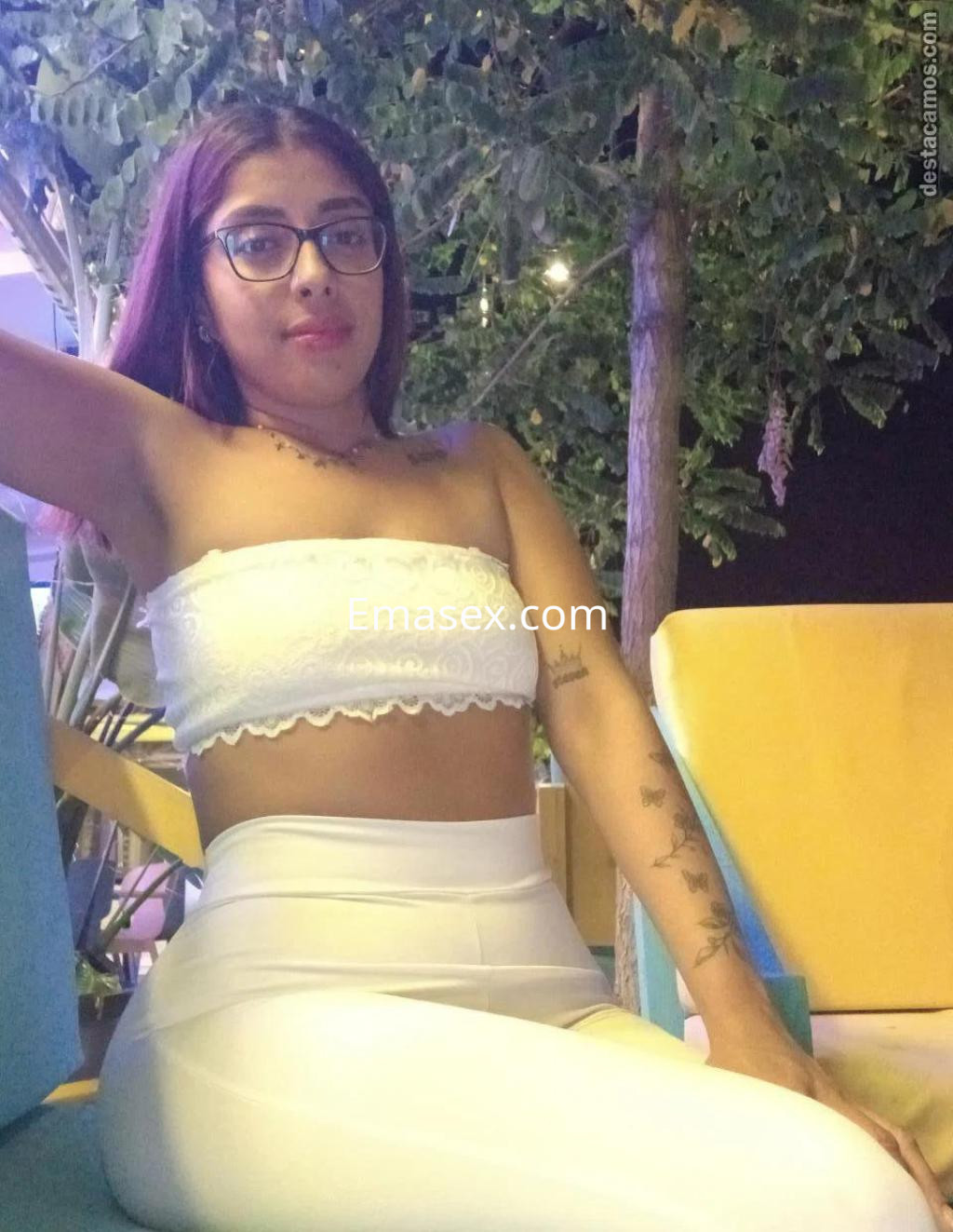 Escort en Zaragoza posa sentada en un sillón, luciendo un top blanco y pantalones ajustados claros, con gafas.