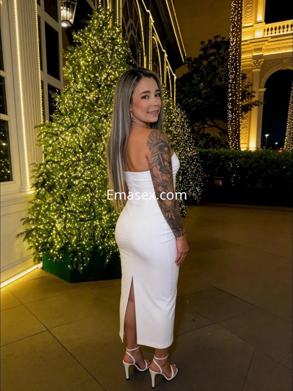 La puta viste un elegante vestido blanco ajustado que resalta su figura, posando ante un árbol iluminado en Zaragoza.