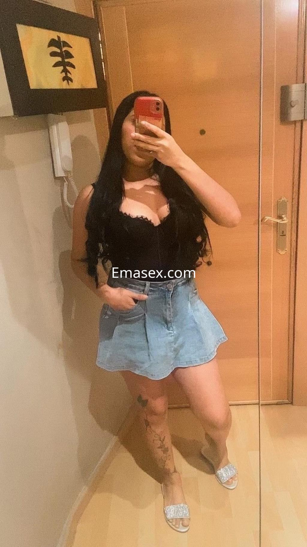 Puta en Zaragoza con cabello largo y oscuro, luciendo un top negro y una falda de mezclilla, posando frente al espejo.