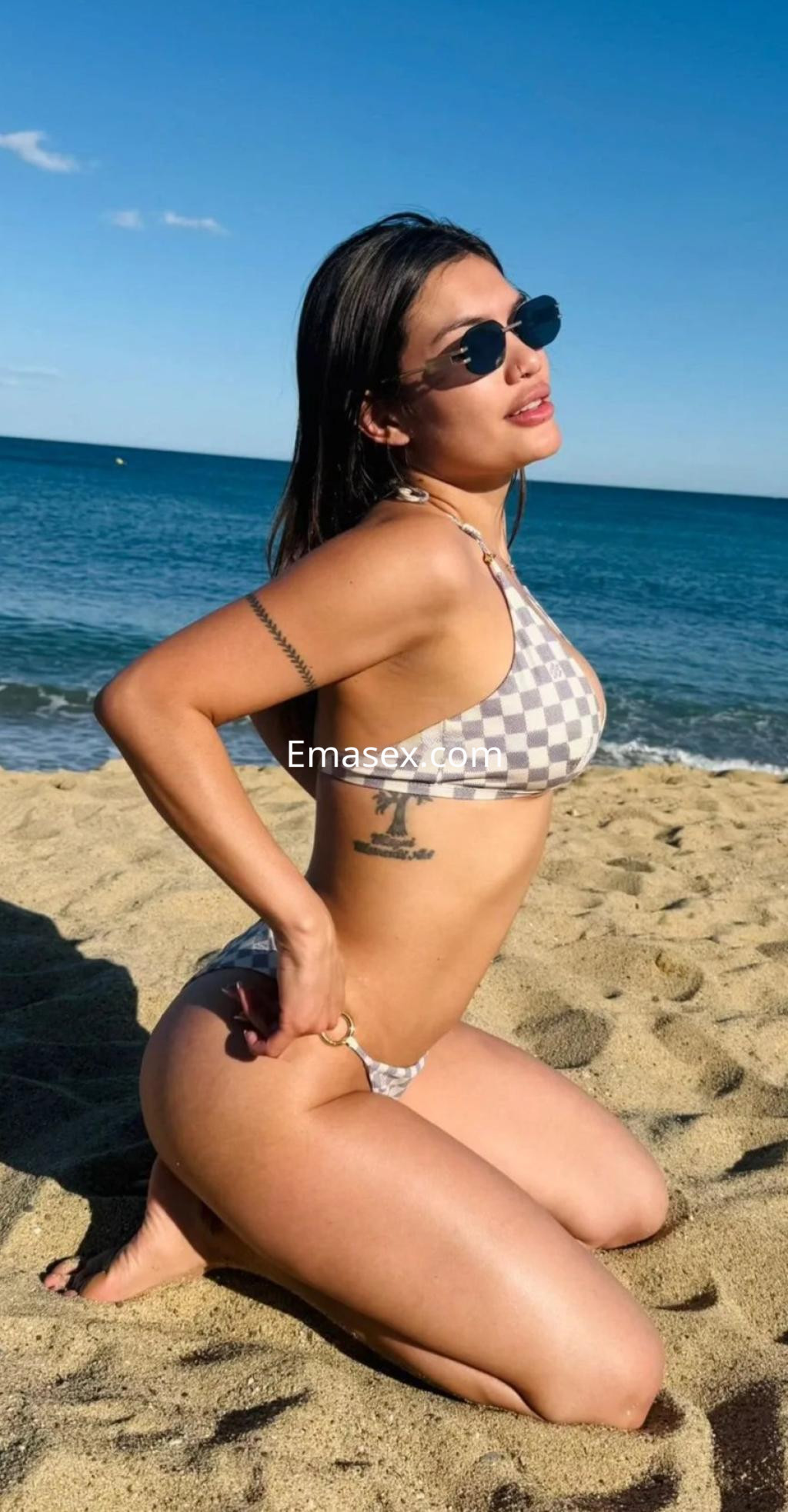 Escort en Palma de Mallorca posando en la playa con un bikini a cuadros y gafas de sol, sobre la arena.