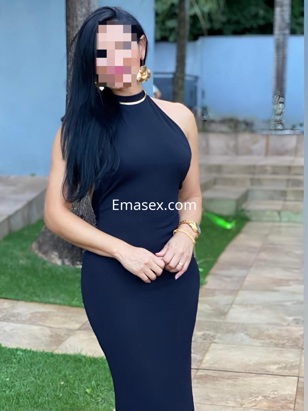 Escort en Bilbao con un vestido negro ajustado, posando de pie en un jardín con césped y árboles al fondo.