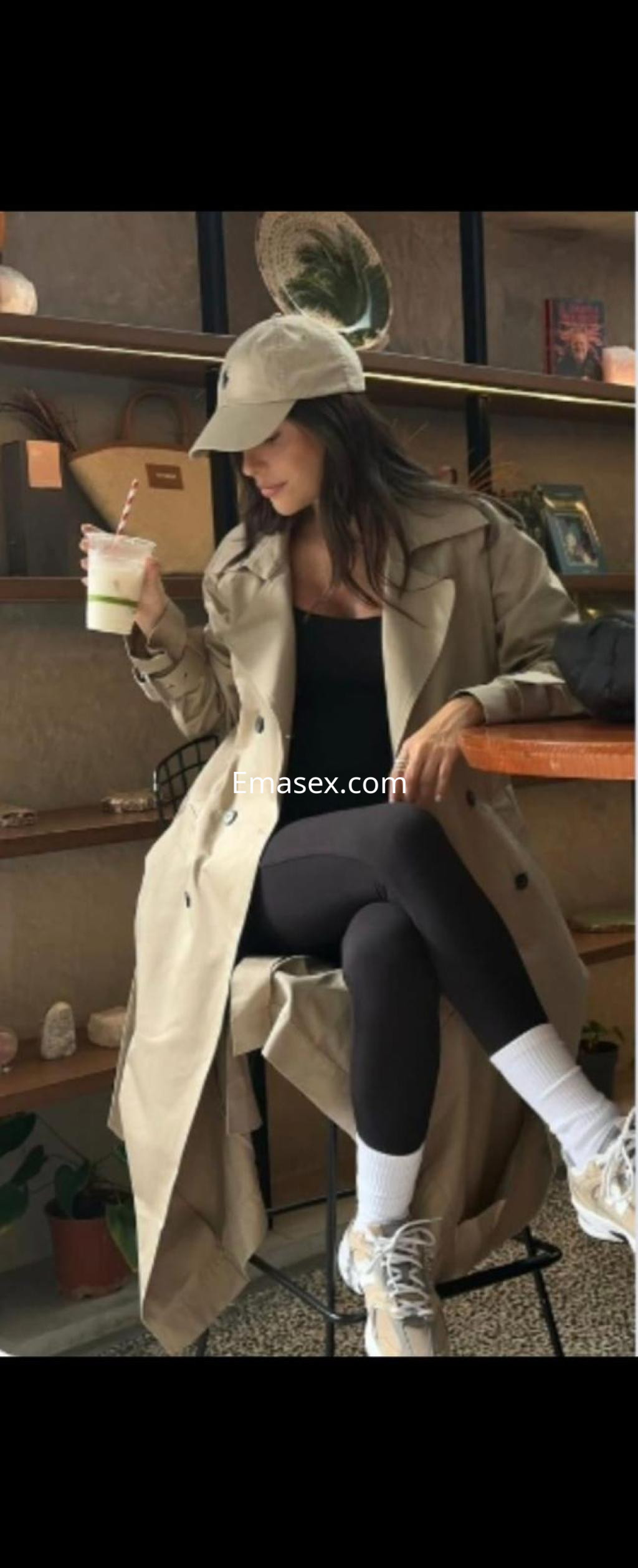 La puta en Bilbao viste un abrigo beige, leggins oscuros y zapatillas, sosteniendo una bebida en un café.