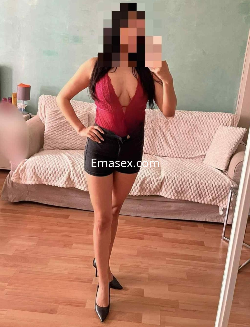 La puta posa en una habitación de Madrid con un body rojo y pantalones cortos negros, luciendo tacones altos.