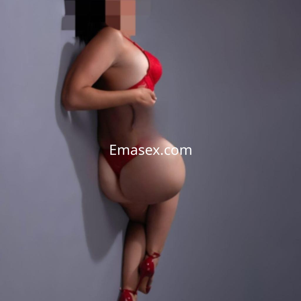 Escort en Barcelona, posando de perfil con lencería roja y tacones, apoyada contra una pared gris.