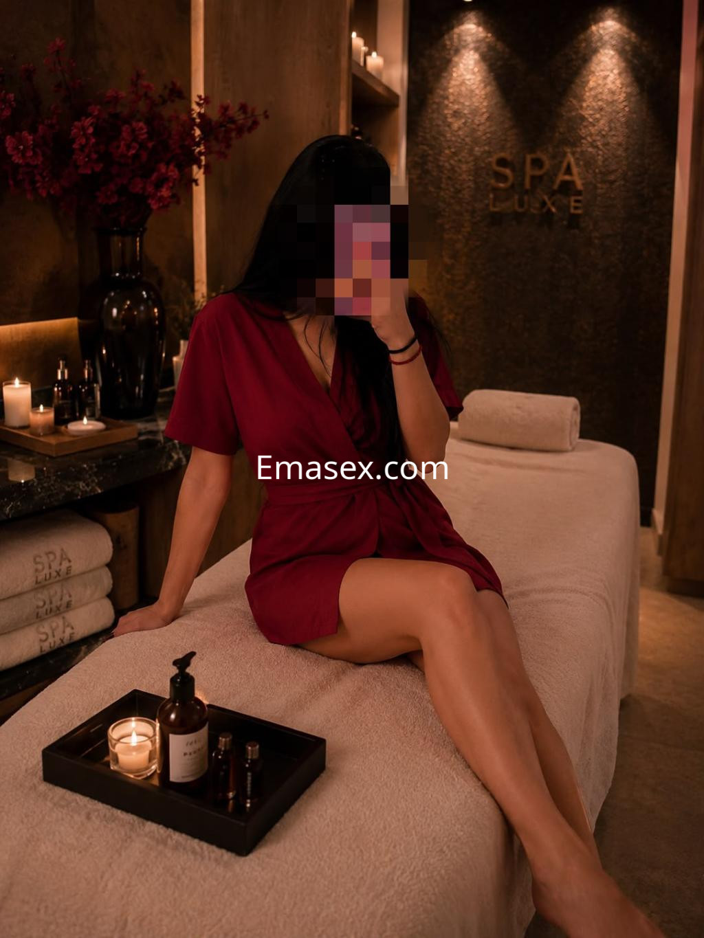 Escort en Gijón, vistiendo una bata roja, sentada en una camilla de spa con velas y aceites a su alrededor.