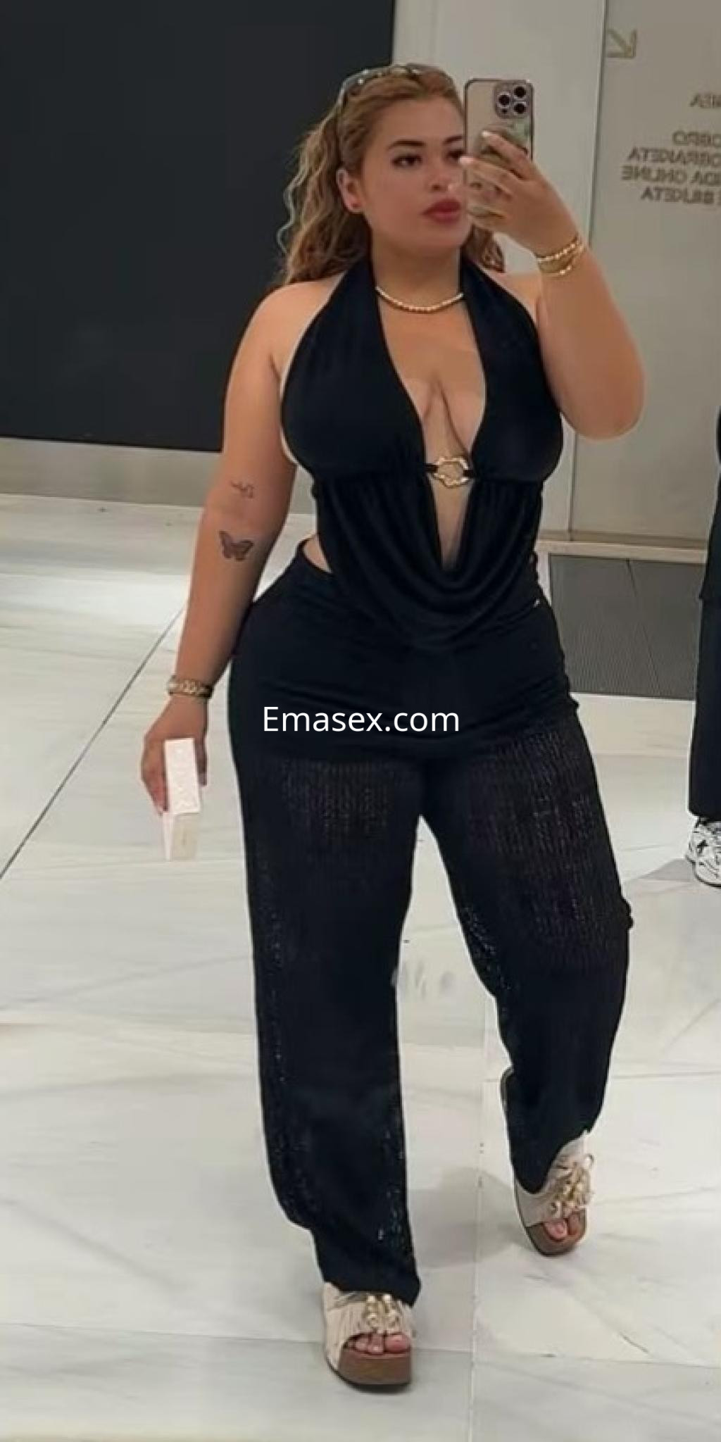 Escort en Bilbao luciendo un conjunto negro con escote y pantalones amplios, posando frente a un espejo.