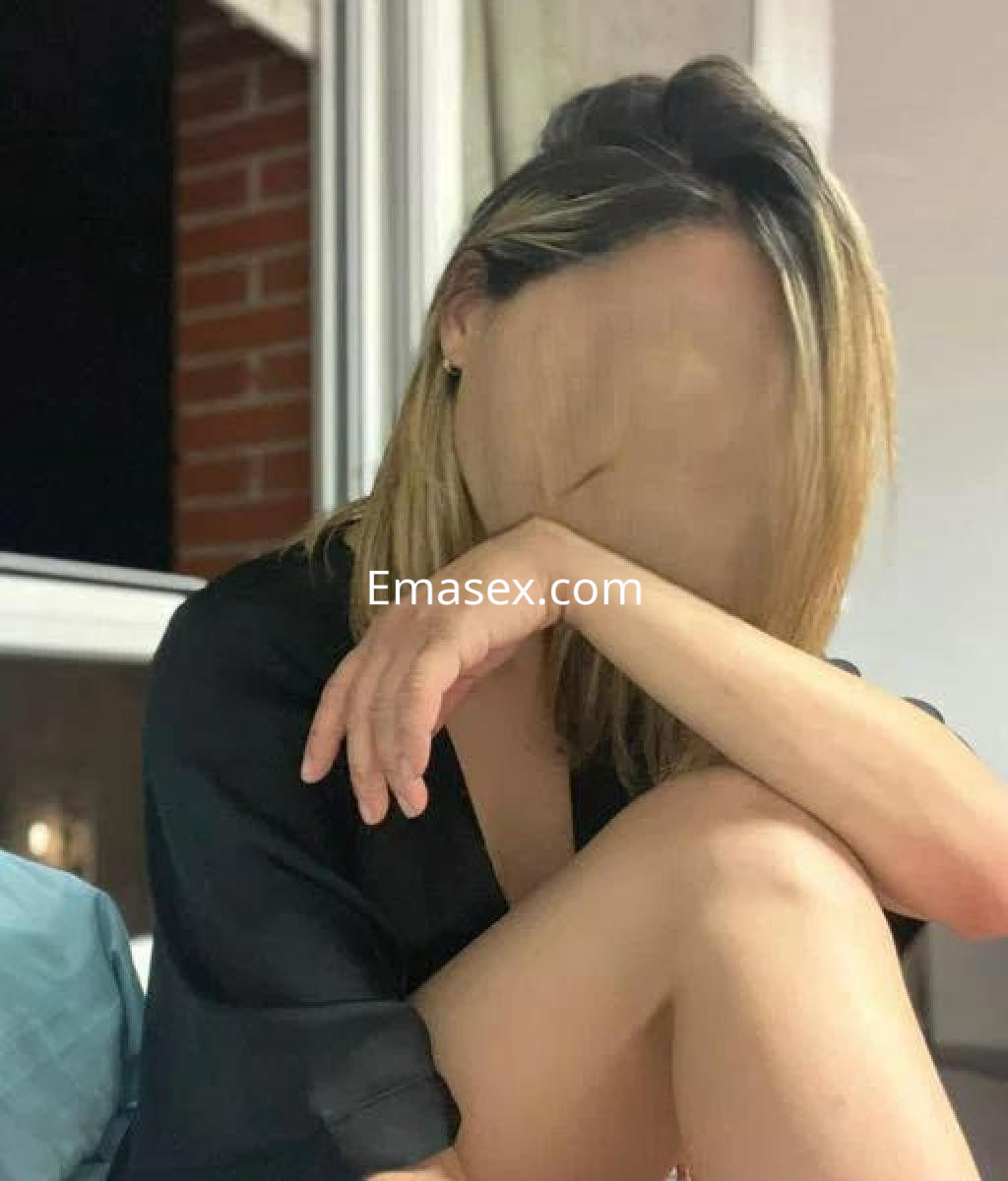 Escort en Barcelona con cabello rubio y lacio, vestida con una blusa negra, sentada en un sofá.