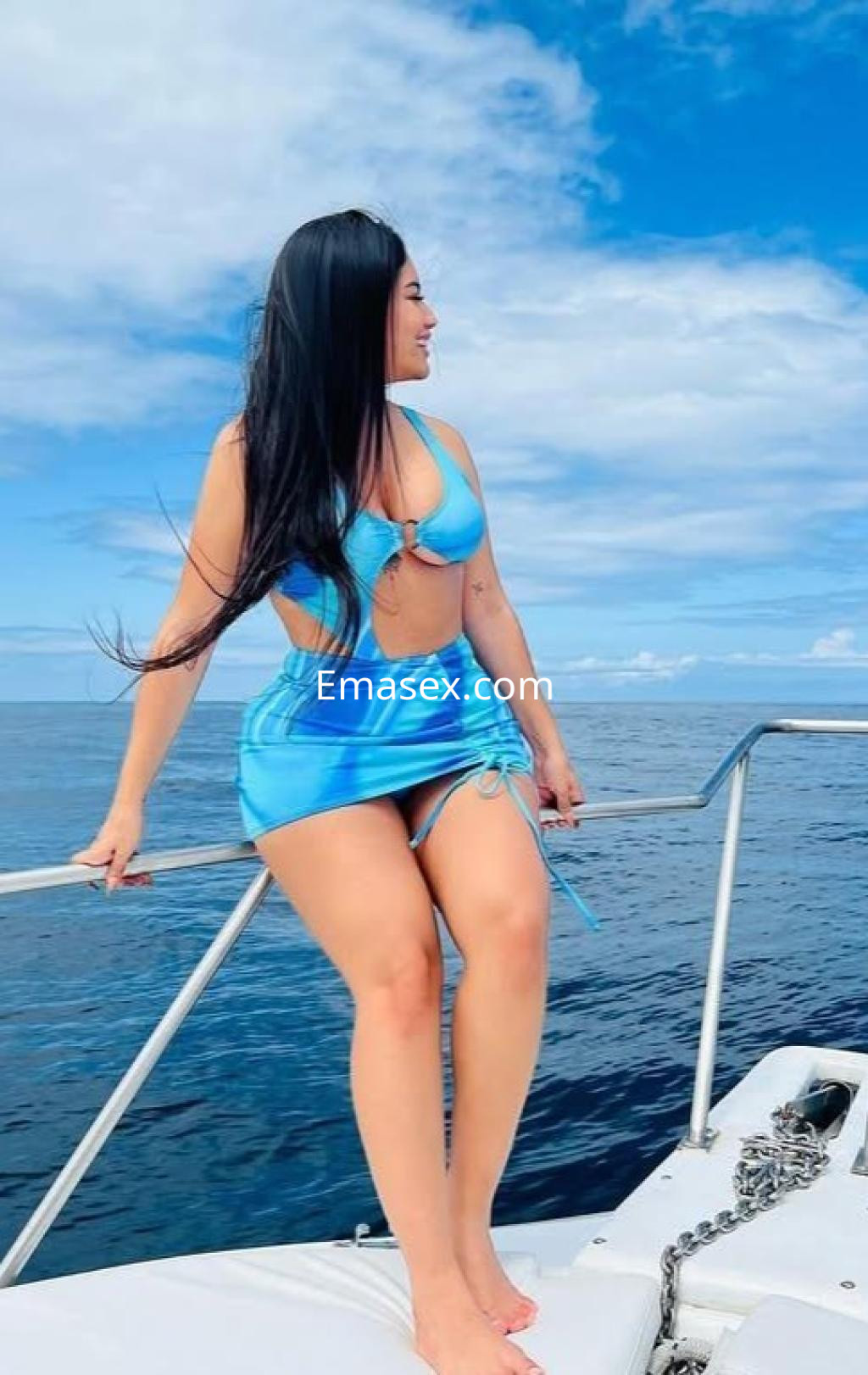 Escort en Zamora, luciendo un bikini azul y una falda a juego, sentada en la borda de un barco.