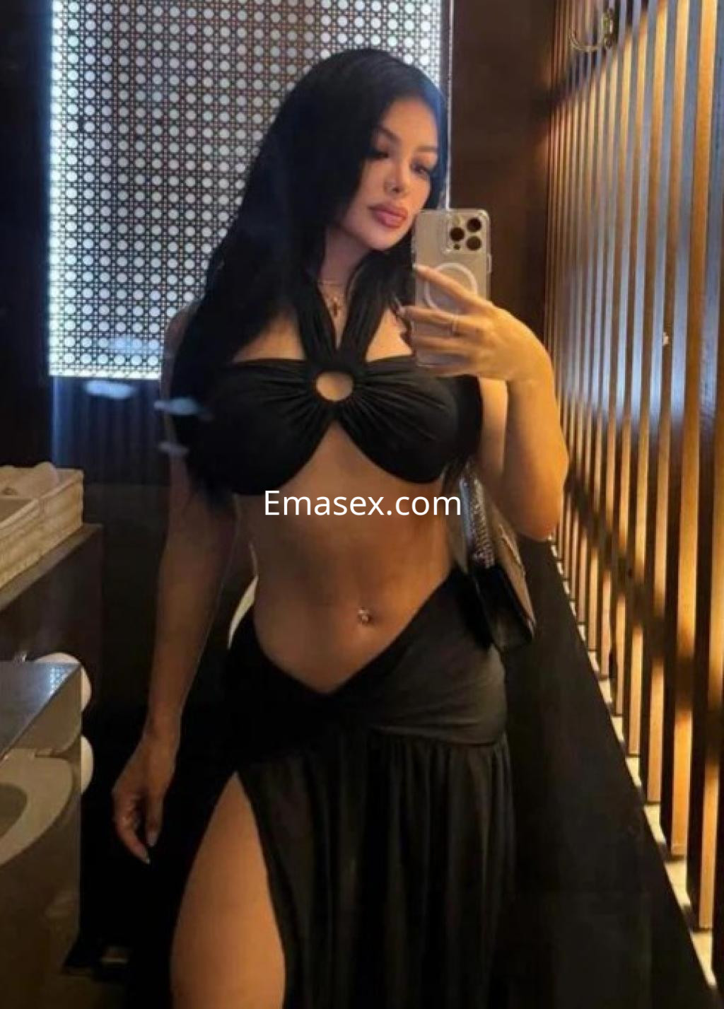 Escort en Valladolid con cabello largo y oscuro, vistiendo un top negro con abertura y falda larga, posando frente a un espejo.