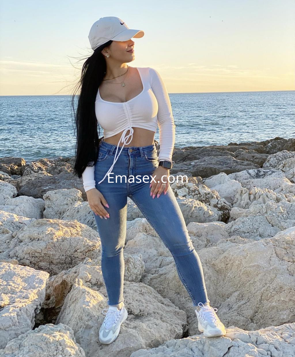 Escort en Vigo con cabello largo y lacio, viste un top blanco y jeans ajustados, posando junto al mar.