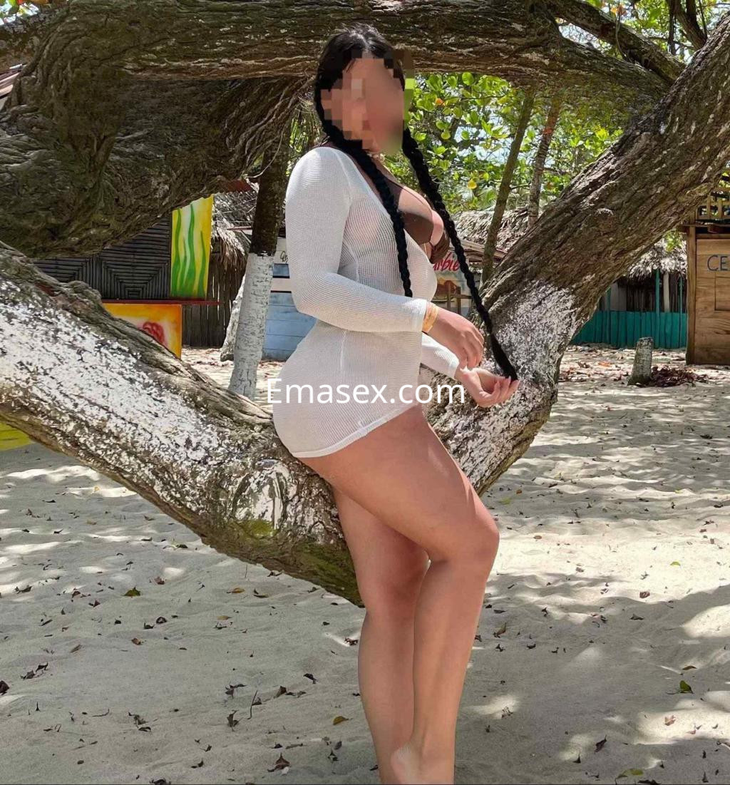Puta de cabello largo y trenzado, viste un conjunto blanco mientras posa junto a un árbol en Barcelona.