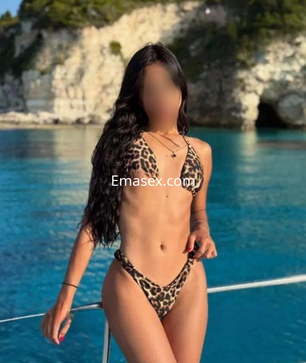 Escort en Barcelona luciendo un bikini de leopardo, con cabello largo y ondulado, frente a un mar azul claro.
