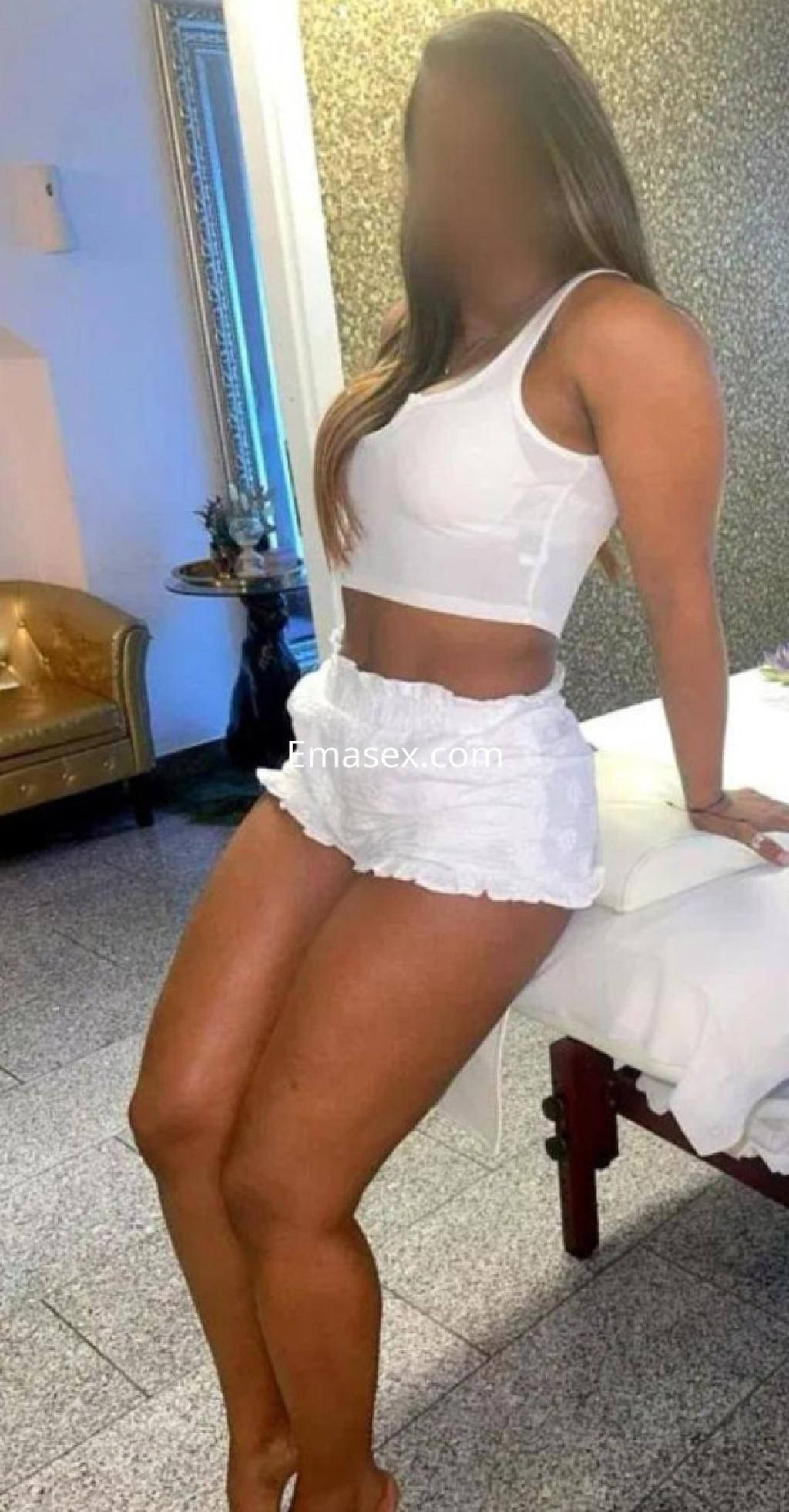 Puta de Granada con top blanco y shorts cortos, posando de pie junto a una cama en un ambiente elegante.