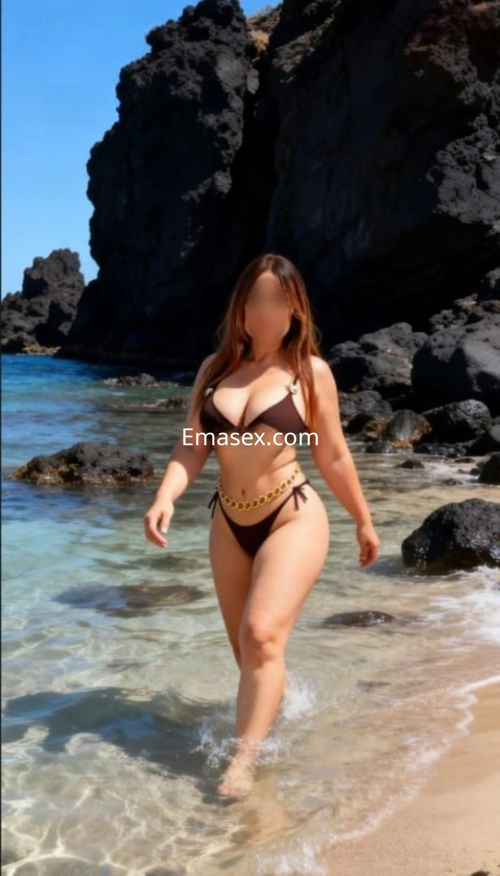 Escort en Madrid caminando en la orilla del mar, con un bikini claro y cabello largo, rodeada de rocas oscuras.