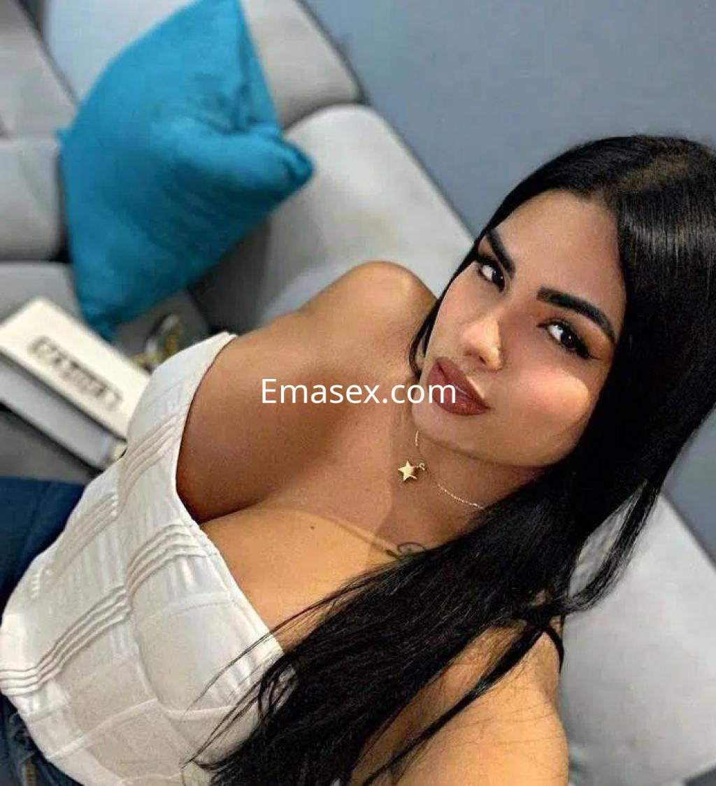 Puta con cabello largo y oscuro, luciendo un top blanco que resalta su figura, en un sofá en Madrid.