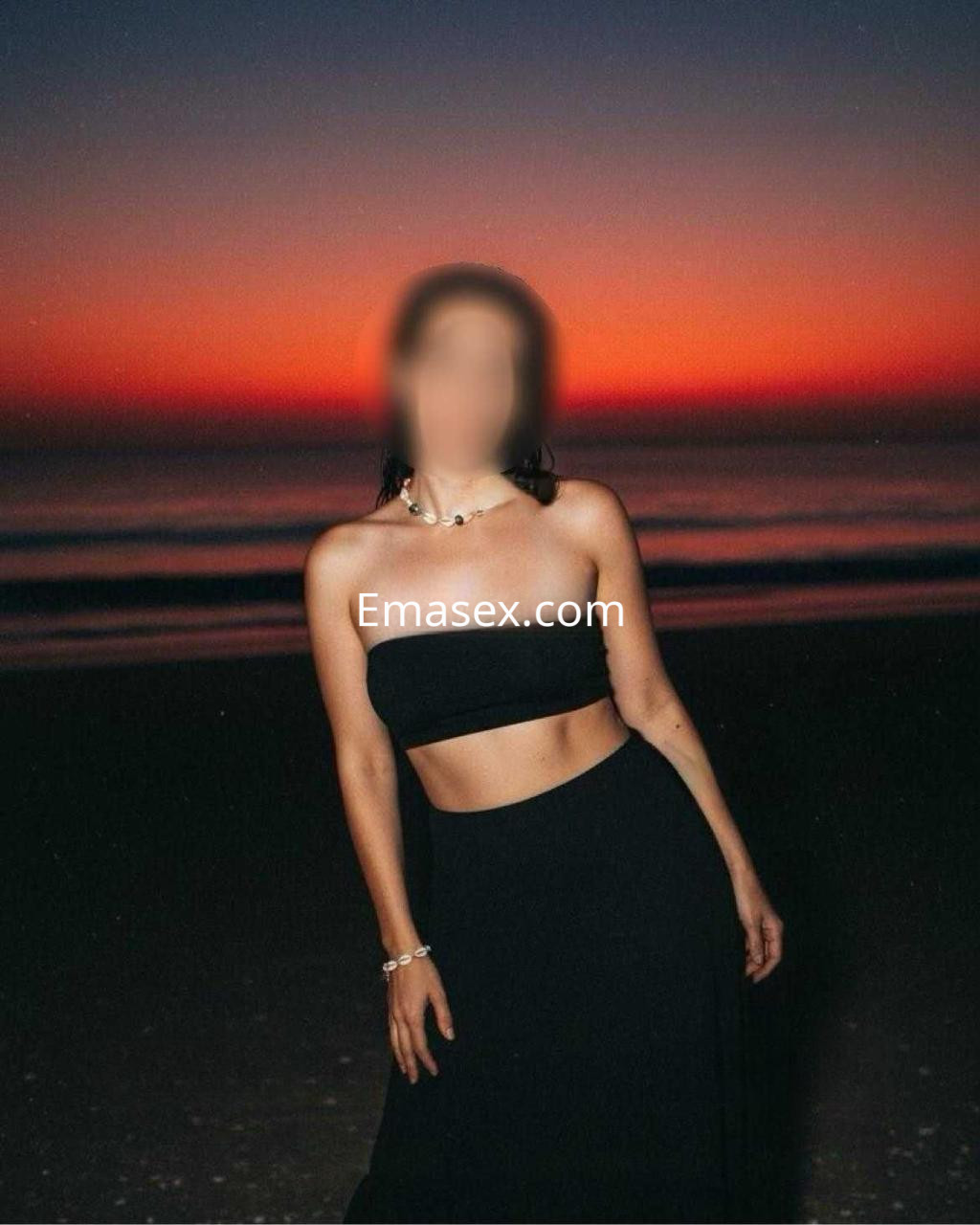Escort en Madrid con top negro sin mangas y falda larga, posando en la playa al atardecer con fondo anaranjado.