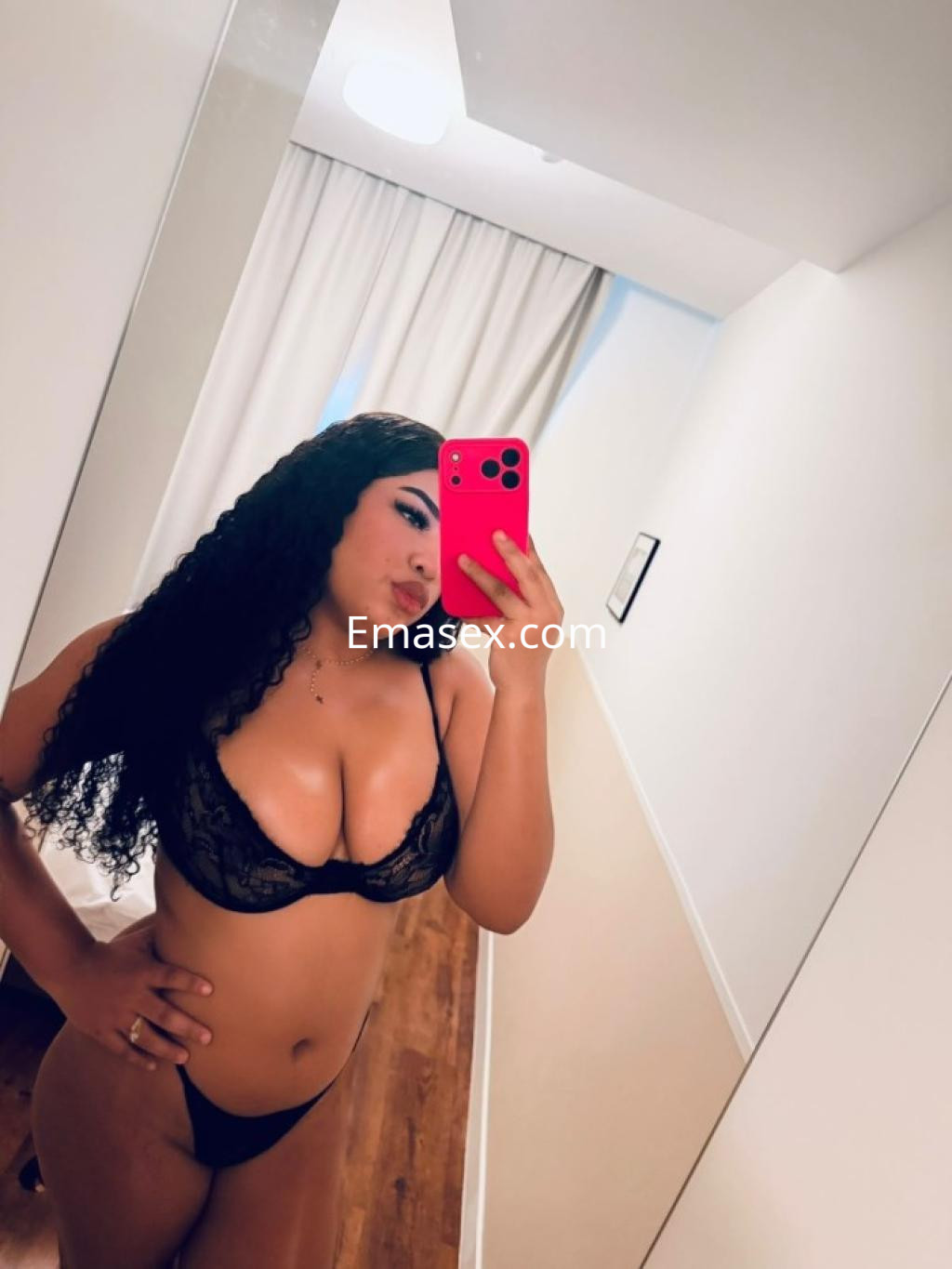 La puta en Tenerife posa en lencería negra, mostrando su figura con cabello rizado y un espejo de fondo.