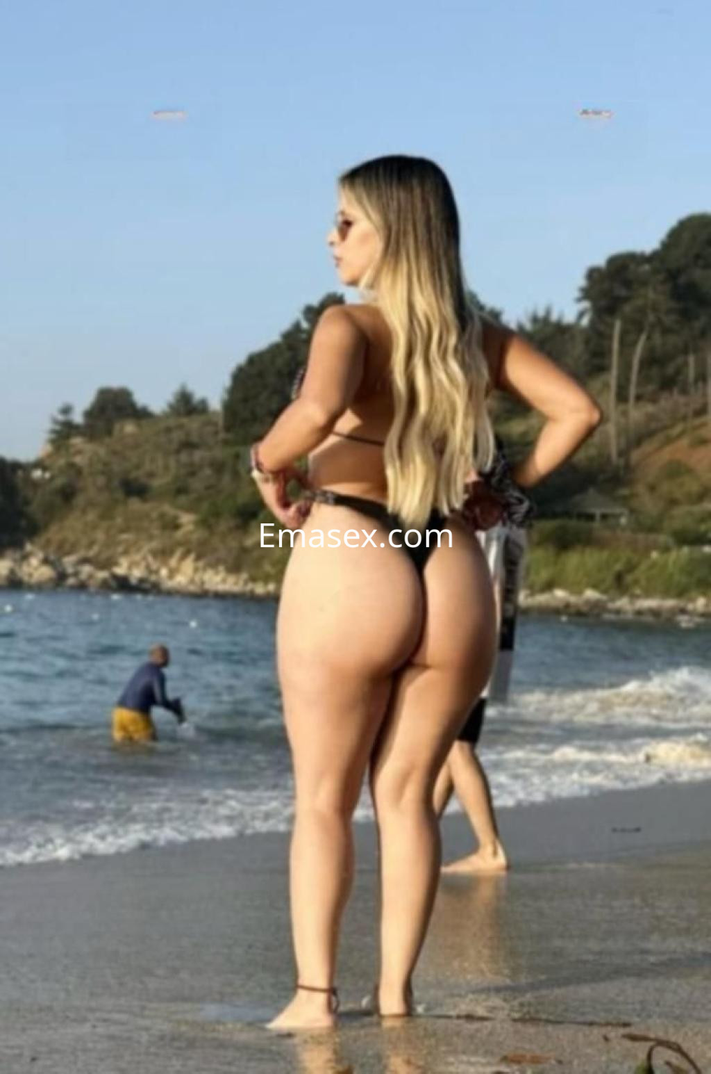 Escort de Zamora en la playa, luciendo un bikini negro y gafas de sol, con el mar de fondo.