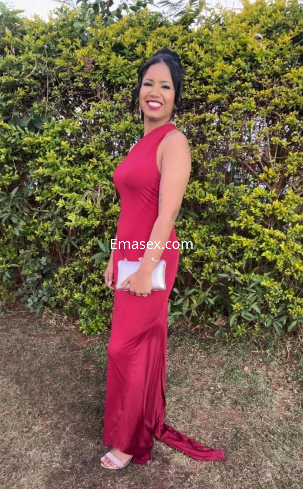 Escort en Irún con vestido largo rojo, de corte ajustado, sonríe mientras se encuentra de pie en un jardín.