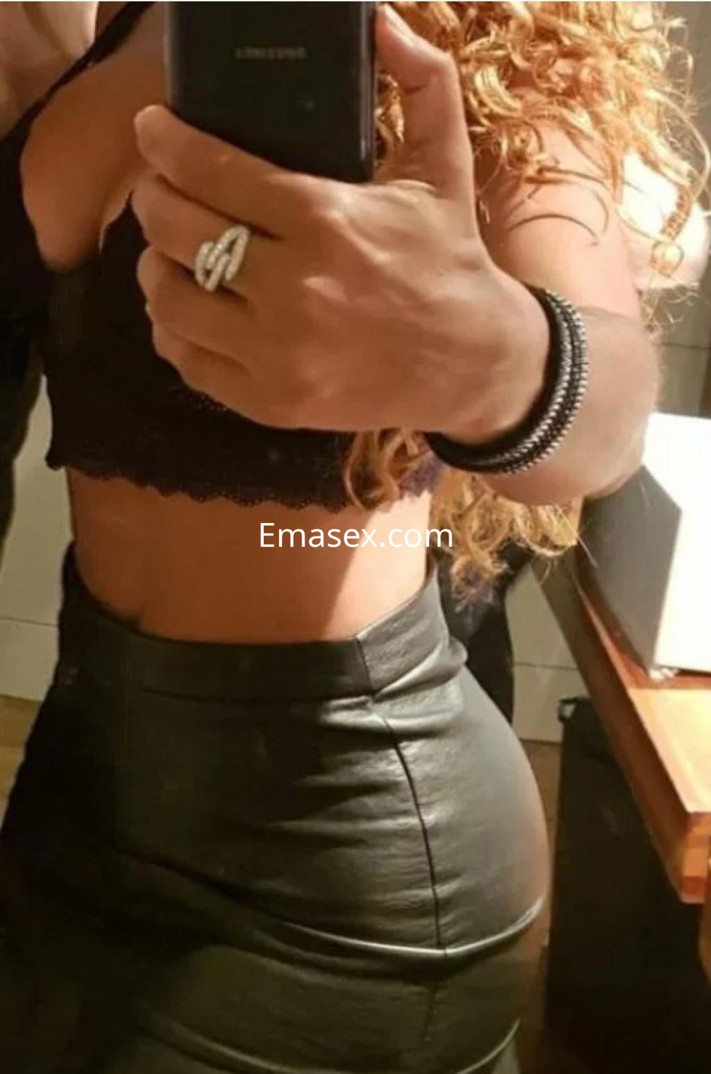 Puta en Vigo con cabello rizado, luciendo un top negro y una falda de cuero ajustada, posando frente al espejo.
