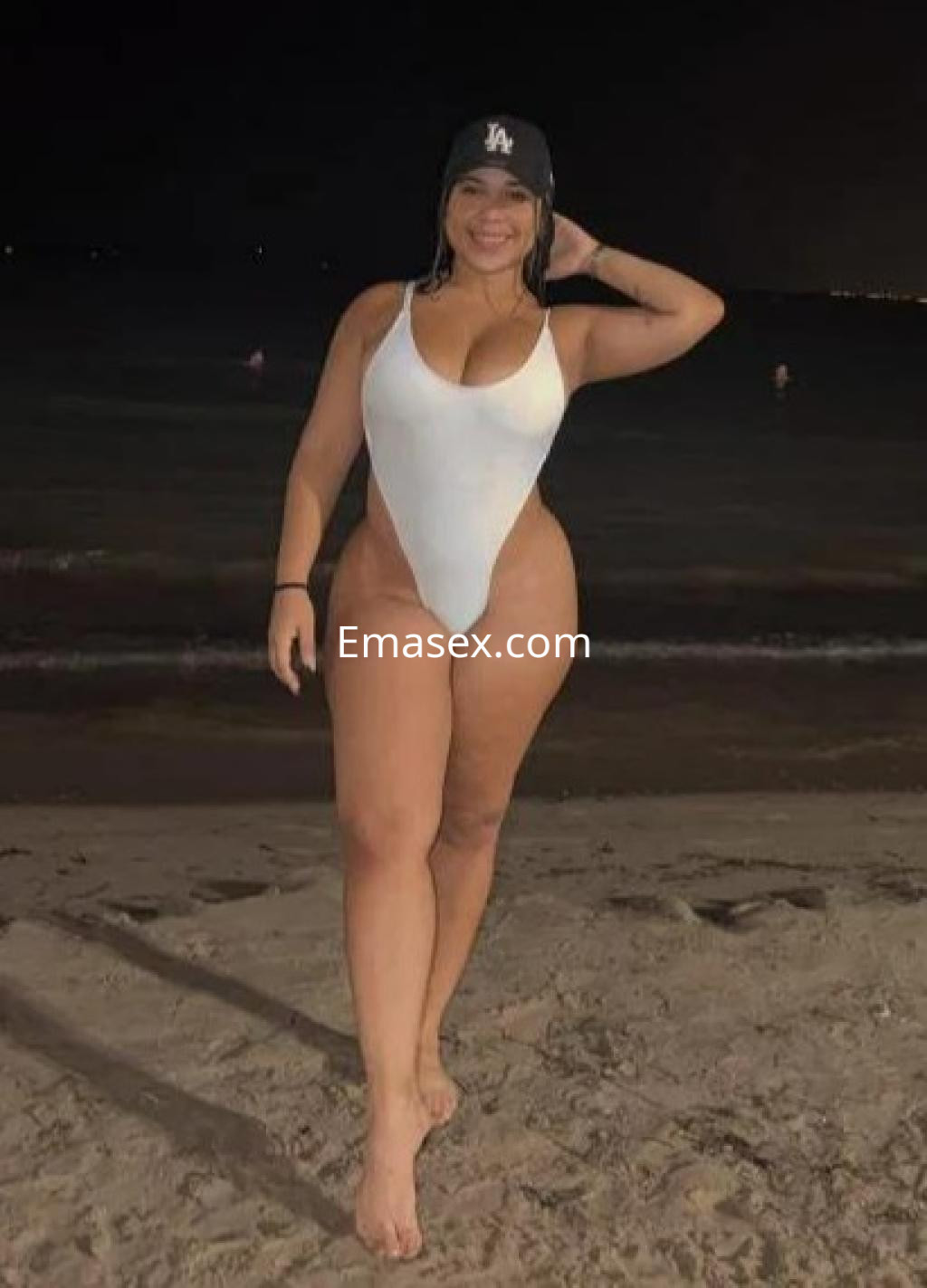 Escort en Madrid luciendo un bañador blanco, de pie en la playa, con una gorra negra y el mar de fondo.