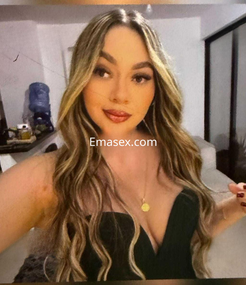 Escort en Madrid con cabello largo y ondulado, luciendo un vestido negro y un collar en un entorno interior.