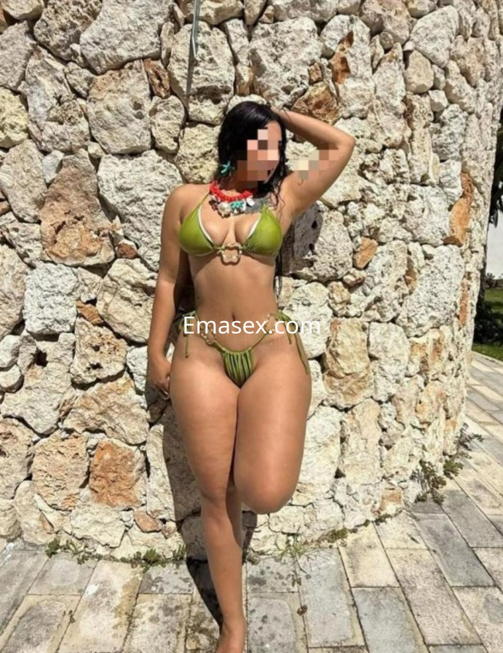 Escort posando con un bikini verde y accesorios coloridos frente a una pared de piedra en Barcelona.