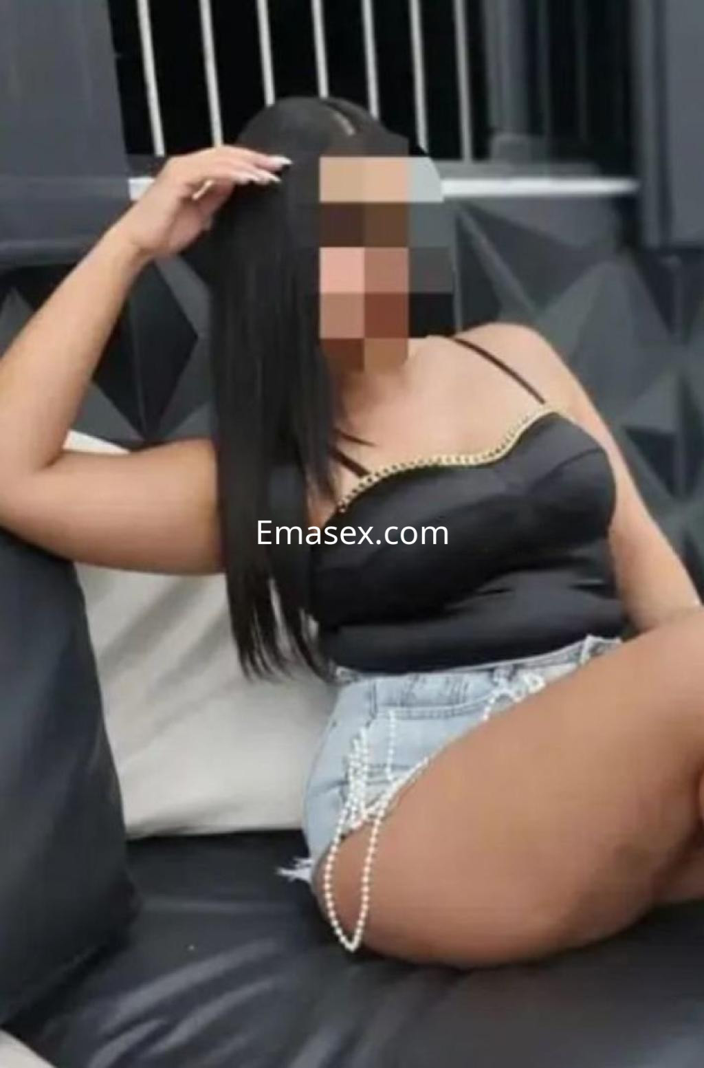 La puta en Murcia lleva una blusa negra sin mangas y shorts de mezclilla, sentada en un sofá moderno.