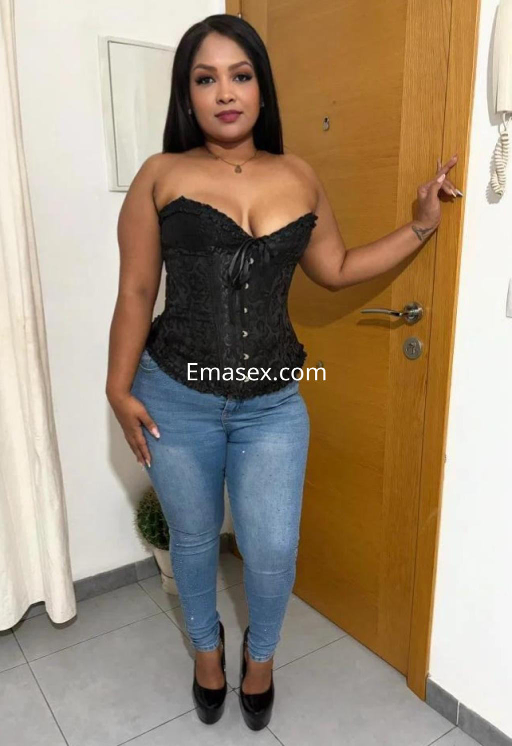 Puta de Cádiz posando en un corsé negro y jeans ajustados, con tacones altos, frente a una puerta de madera.
