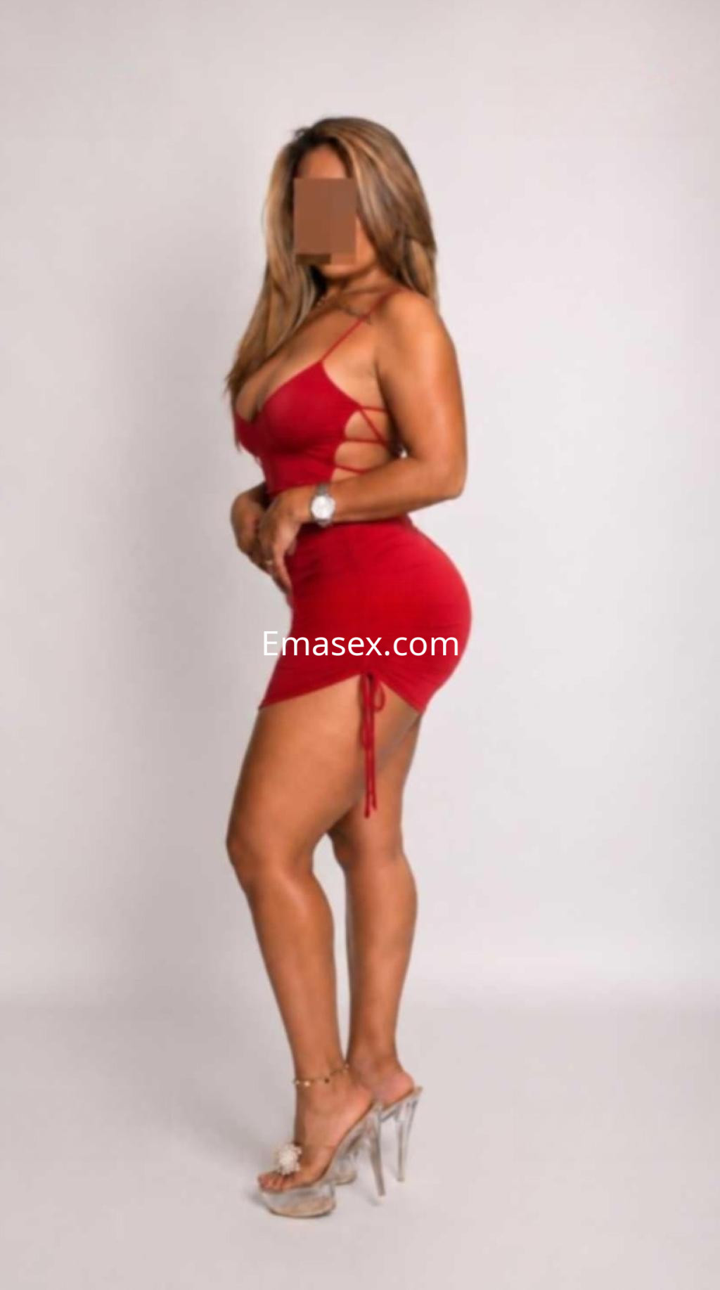 Escort en Ibiza luciendo un vestido rojo ajustado con tirantes y sandalias de tacón, de pie en un fondo neutro.