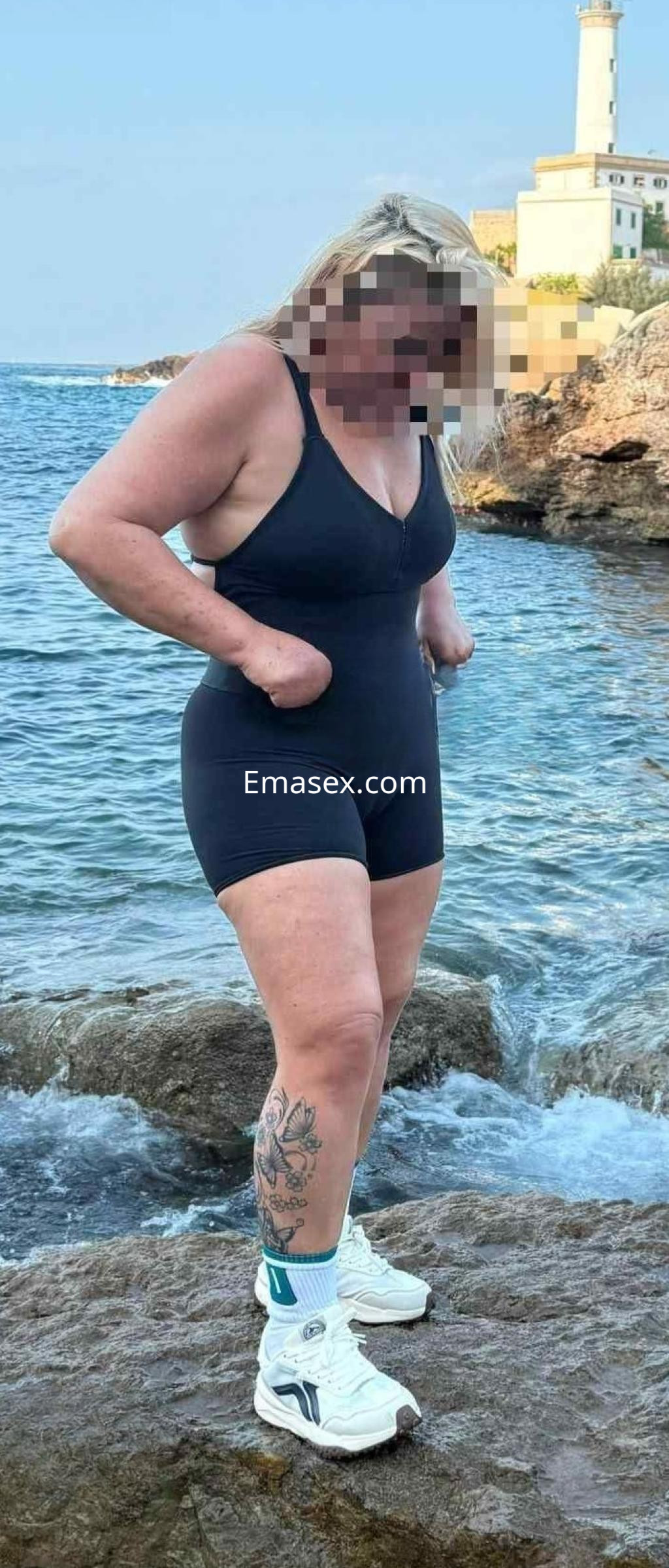 Escort en Ibiza con cabello rubio, usando un traje de baño negro y zapatillas deportivas, posando junto al mar.