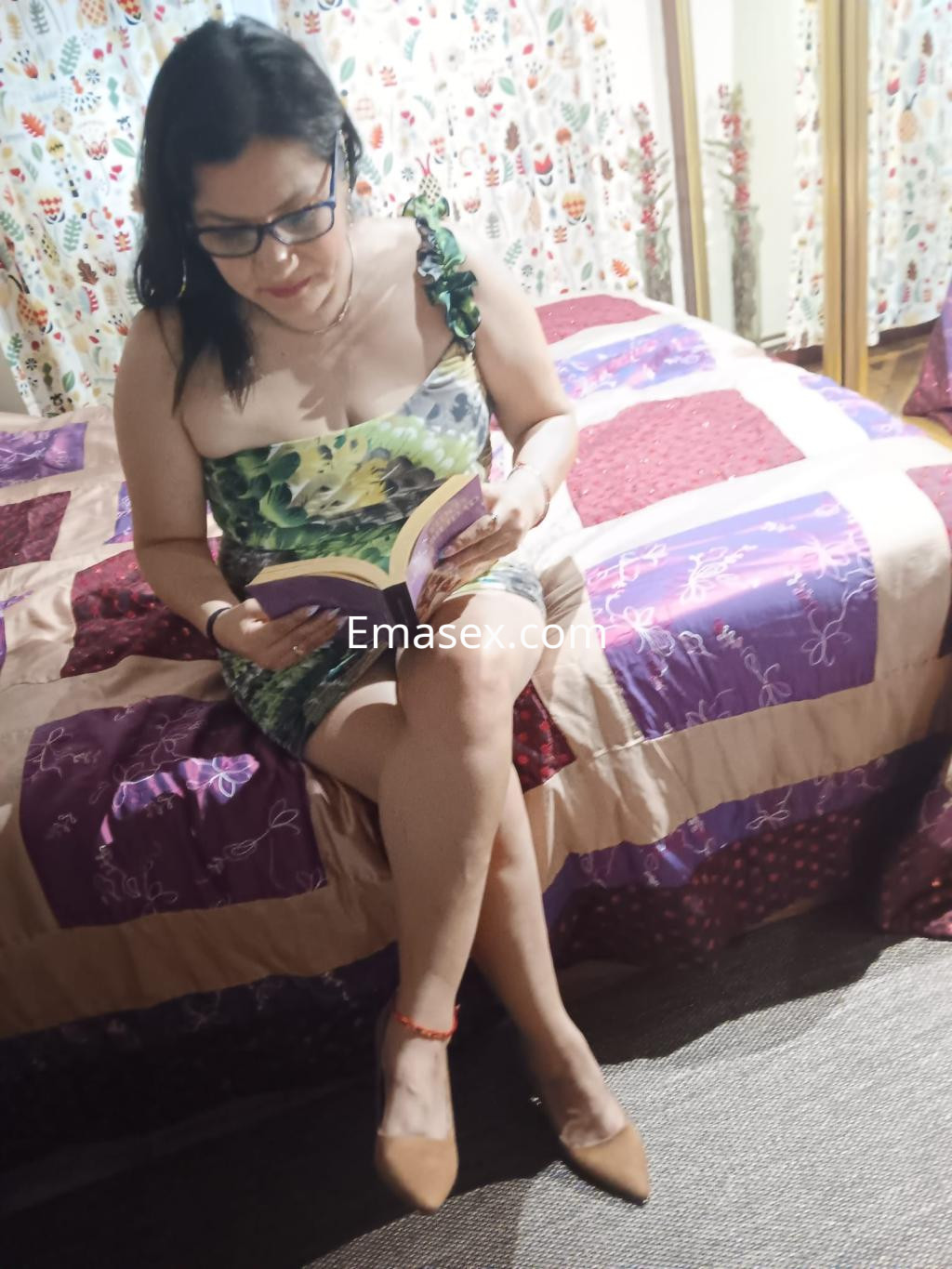 Escort en Santander, sentada en una cama con sábanas de colores, leyendo un libro y luciendo un vestido de tirante.