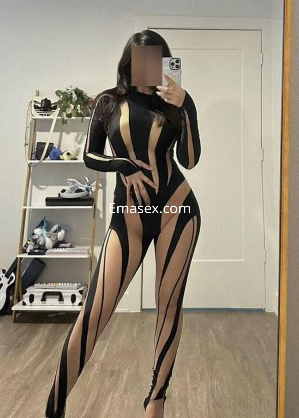 Puta en Barcelona viste un bodysuit negro con líneas transparentes, posando frente a un espejo en un ambiente moderno.