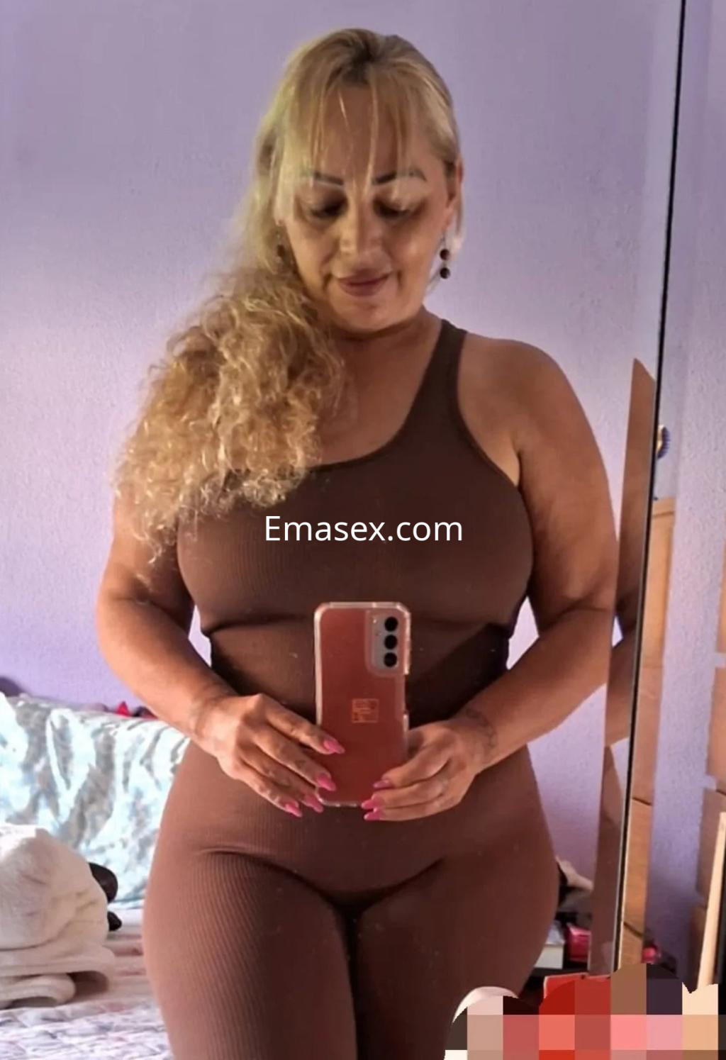 Escort en Oviedo con cabello rubio rizado, viste un mono marrón y se toma una selfie frente a un espejo.