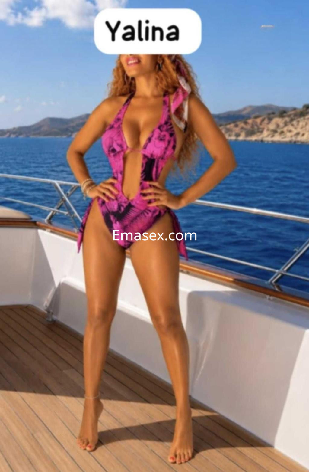 Escort en Valladolid vistiendo un bañador rosa con estampado, de pie en un yate frente al mar.