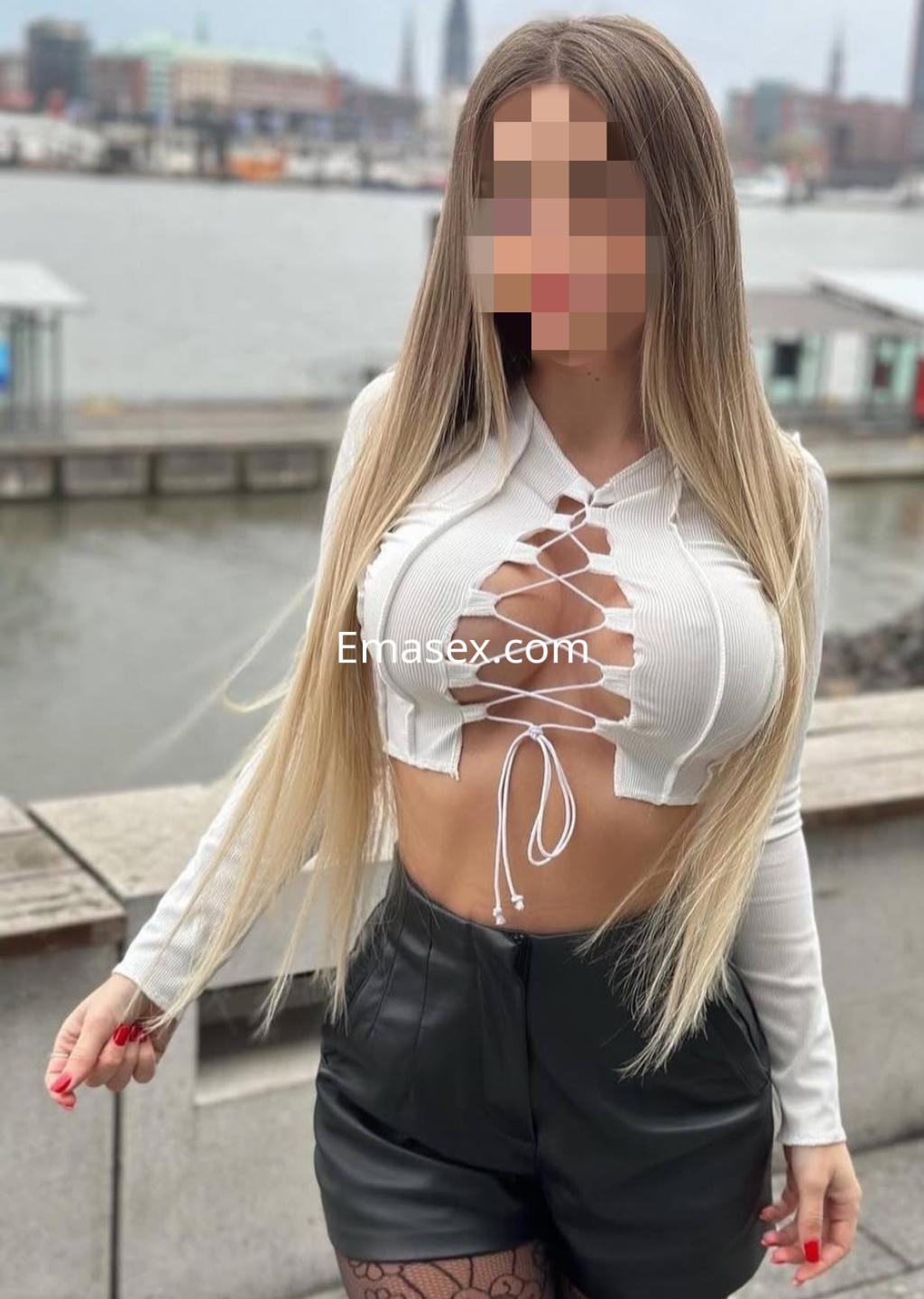 Escort en Santander posando junto al agua, vistiendo un top blanco con detalles de encaje y pantalones cortos negros.